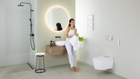 Žena naslonjena na umivaonik u kupaonici s tuš WC-om Geberit AquaClean Sela te umivaonikom i namještajem Geberit VariForm